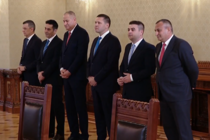Six men in dark suits stand in a row in a formal room, hands clasped, facing forward near ornate wooden chairs and patterned wallpaper.