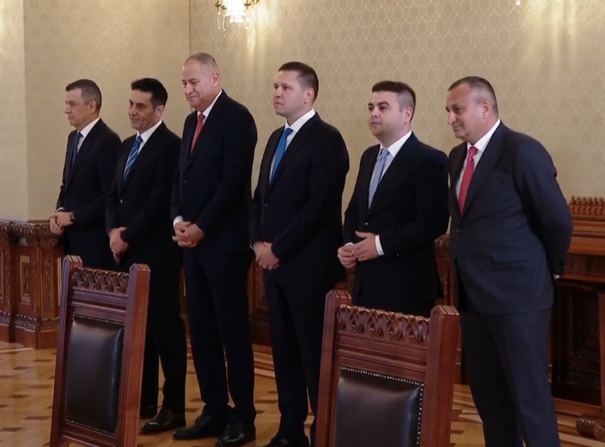 Six men in dark suits stand in a row in a formal room, hands clasped, facing forward near ornate wooden chairs and patterned wallpaper.