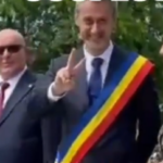 Man wearing a tricolor sash (blue, yellow, red) raises two fingers in a peace-like gesture at an outdoor event, flanked by a suited man with sunglasses and a woman.