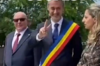 Man wearing a tricolor sash (blue, yellow, red) raises two fingers in a peace-like gesture at an outdoor event, flanked by a suited man with sunglasses and a woman.