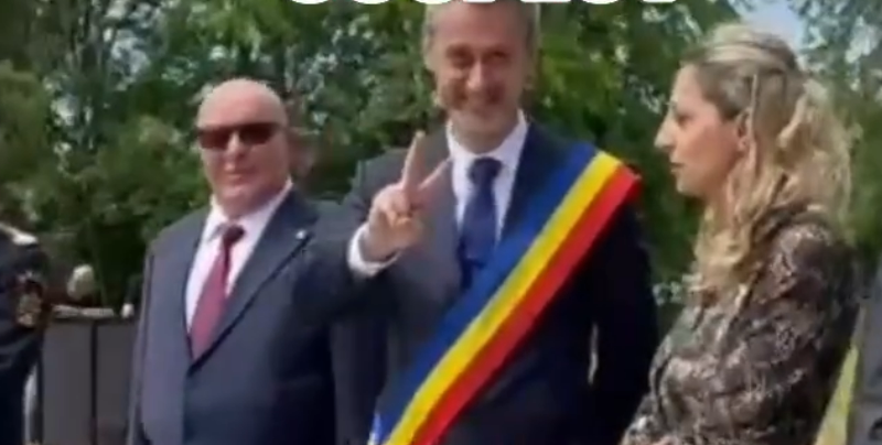 Man wearing a tricolor sash (blue, yellow, red) raises two fingers in a peace-like gesture at an outdoor event, flanked by a suited man with sunglasses and a woman.