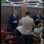 Two barbers in blue suits work on a customer seated in a barber chair inside a modern shop with geometric ceiling lights and wooden dividers.
