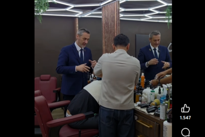 Two barbers in blue suits work on a customer seated in a barber chair inside a modern shop with geometric ceiling lights and wooden dividers.