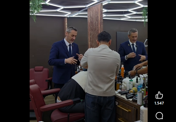 Two barbers in blue suits work on a customer seated in a barber chair inside a modern shop with geometric ceiling lights and wooden dividers.