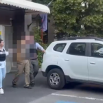 Officer in a dark vest stands near a white SUV as two blurred individuals approach in a parking lot outside a building with a small awning.
