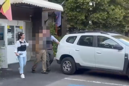 Officer in a dark vest stands near a white SUV as two blurred individuals approach in a parking lot outside a building with a small awning.