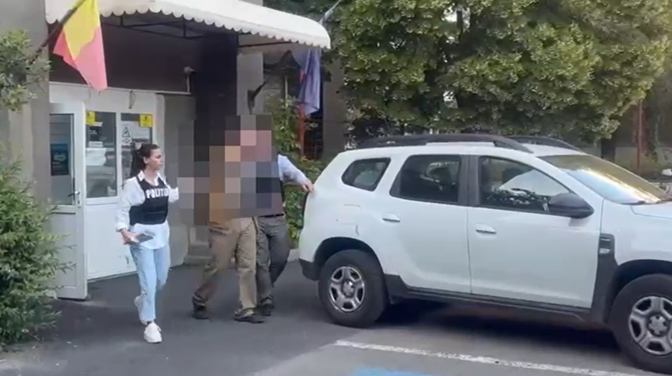 Officer in a dark vest stands near a white SUV as two blurred individuals approach in a parking lot outside a building with a small awning.