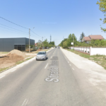 Straight two-lane street with a car ahead, construction pile on the left, modern buildings, white wall and trees on the right under a clear blue sky. It shows Strada ... painted on the road.