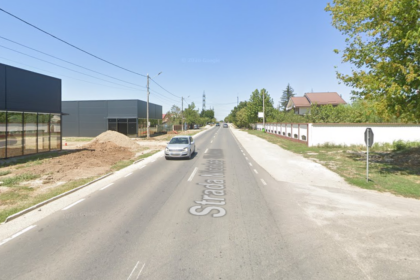 Straight two-lane street with a car ahead, construction pile on the left, modern buildings, white wall and trees on the right under a clear blue sky. It shows Strada ... painted on the road.
