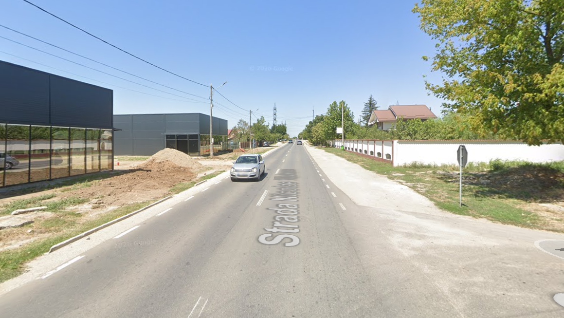 Screenshot 2026-05-29 103316 - Jurnalul Olteniei – Știri din Craiova și Oltenia Straight two-lane street with a car ahead, construction pile on the left, modern buildings, white wall and trees on the right under a clear blue sky. It shows Strada ... painted on the road.