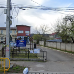 Entrance to a local government office complex with a blue sign, gated yard, and street view; residential buildings and a small park to the left, overhead power lines visible.