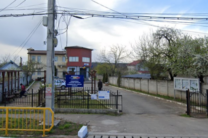 Entrance to a local government office complex with a blue sign, gated yard, and street view; residential buildings and a small park to the left, overhead power lines visible.