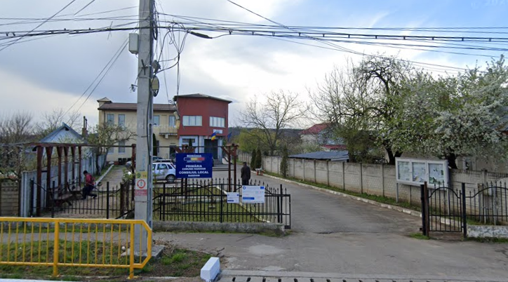 Entrance to a local government office complex with a blue sign, gated yard, and street view; residential buildings and a small park to the left, overhead power lines visible.
