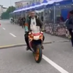 Rider on a sport bike with a passenger on the back, approaching the camera on a city street lined with spectators and barricades.