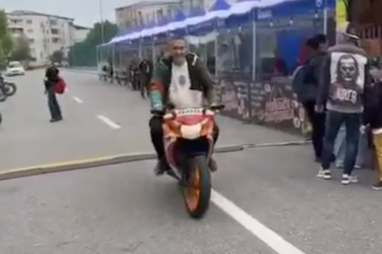 Rider on a sport bike with a passenger on the back, approaching the camera on a city street lined with spectators and barricades.