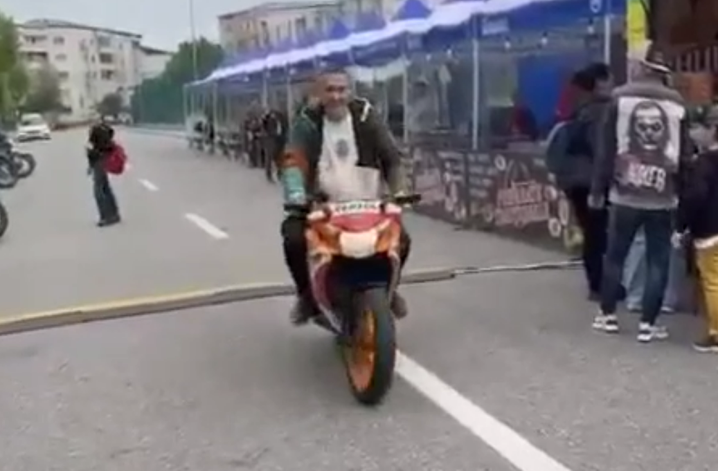 Rider on a sport bike with a passenger on the back, approaching the camera on a city street lined with spectators and barricades.
