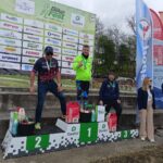 Three race winners stand on a podium at a bike festival, wearing medals; first place in a bright neon green jacket, second in a black-pink kit, third in a blue outfit, with prizes on the steps and sponsor banners behind them.
