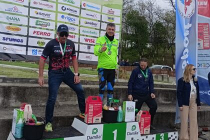 Three race winners stand on a podium at a bike festival, wearing medals; first place in a bright neon green jacket, second in a black-pink kit, third in a blue outfit, with prizes on the steps and sponsor banners behind them.