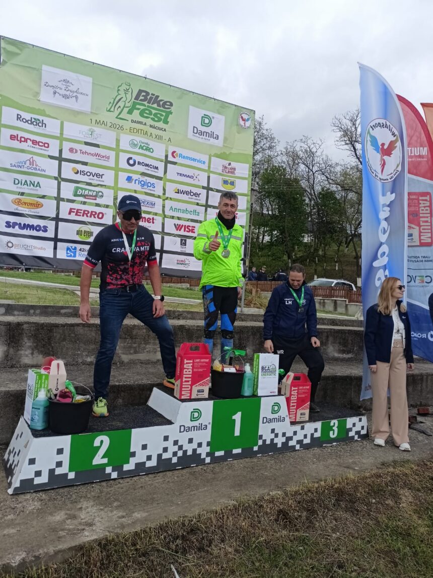 Three race winners stand on a podium at a bike festival, wearing medals; first place in a bright neon green jacket, second in a black-pink kit, third in a blue outfit, with prizes on the steps and sponsor banners behind them.