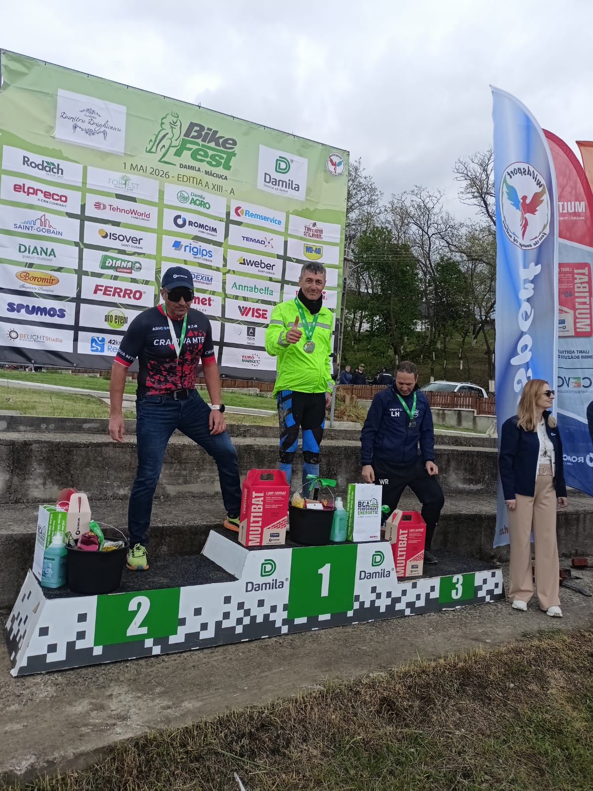 Three race winners stand on a podium at a bike festival, wearing medals; first place in a bright neon green jacket, second in a black-pink kit, third in a blue outfit, with prizes on the steps and sponsor banners behind them.