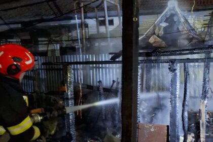 Firefighter in red helmet using a hose to spray water on a burning, collapsed structure at night.