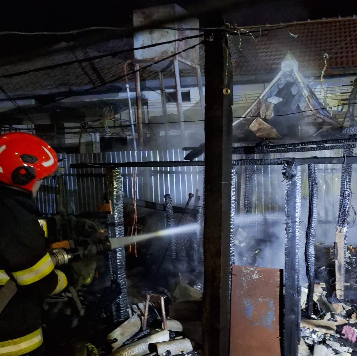 Firefighter in red helmet using a hose to spray water on a burning, collapsed structure at night.