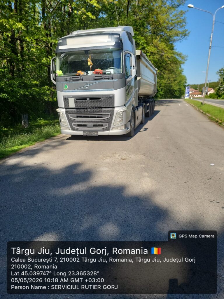 Front view of a large silver semi-truck parked on a rural road with green trees; on-screen GPS overlay shows Târgu Jiu, Romania and coordinates.