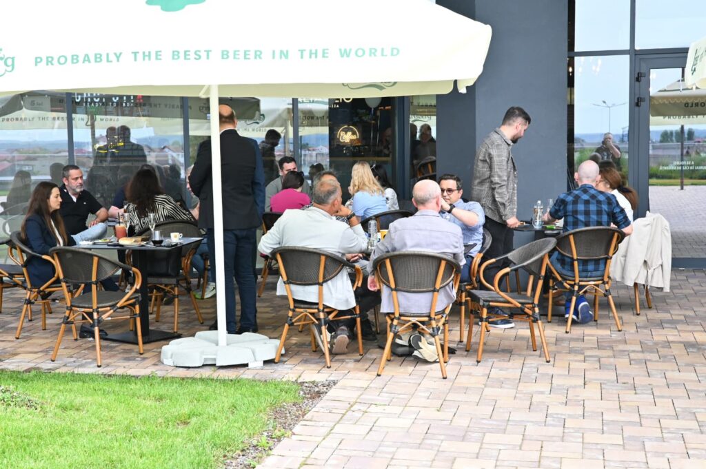 Group of people dining at an outdoor cafe under a large white umbrella on a brick patio, talking and eating.