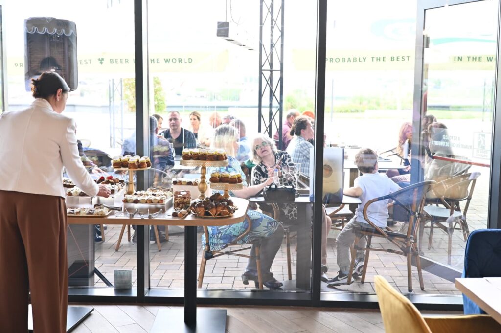 Waiter in a light blazer arranges a dessert display of cupcakes and pastries inside a glass-walled cafe as guests sit at outdoor tables beyond the window.