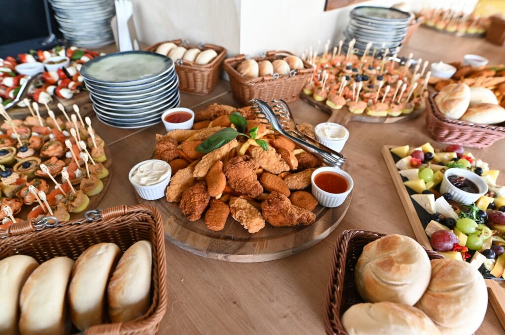 Assorted finger foods on a buffet table: fried chicken bites, bread rolls, fruit and cheese platter, and skewers with dips in small bowls on a wooden board.