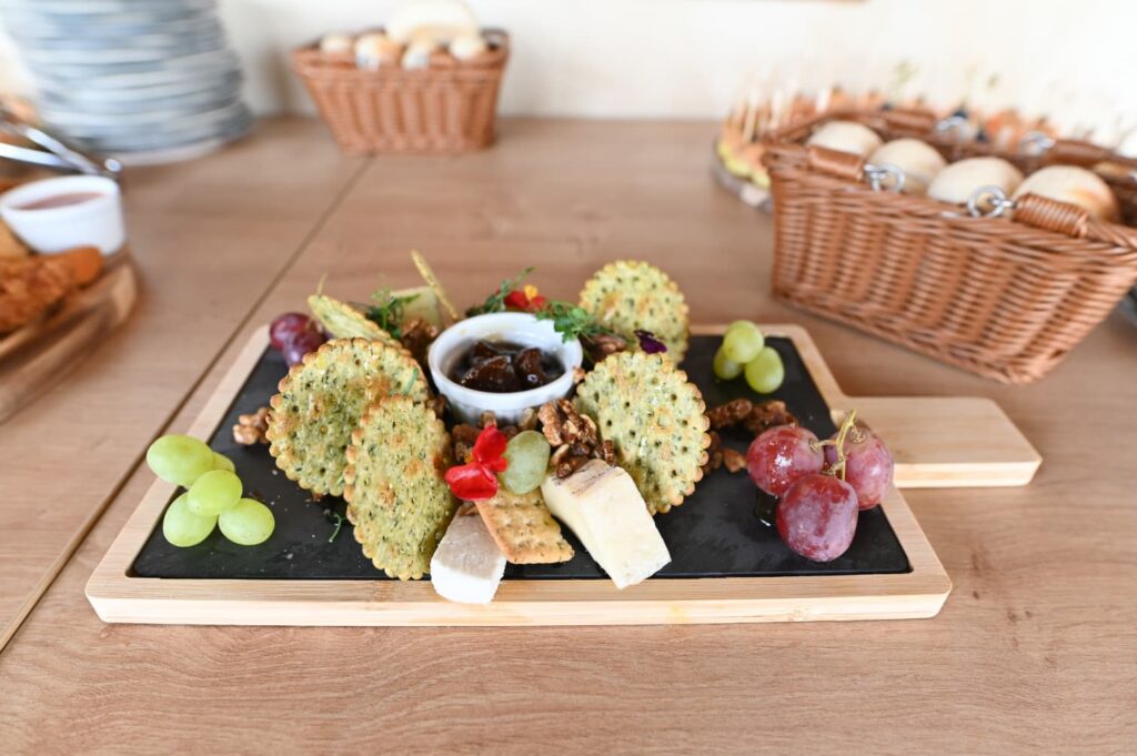 Cheese and cracker platter on a wooden board with grapes, walnuts, and a small bowl of olives in the center