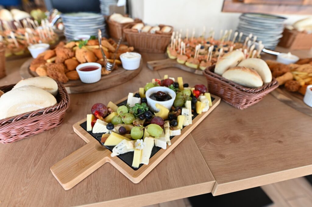 Cheese board with grapes, assorted cheeses, fruit chunks, and a small dip cup on a wooden serving board, on a table with bread baskets in the background.