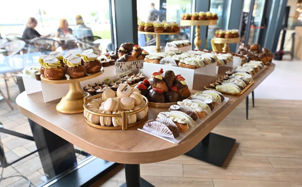 Assorted cupcakes, macarons, and pastries arranged on a wooden dessert table by a glass-walled area of a cafe or event space.