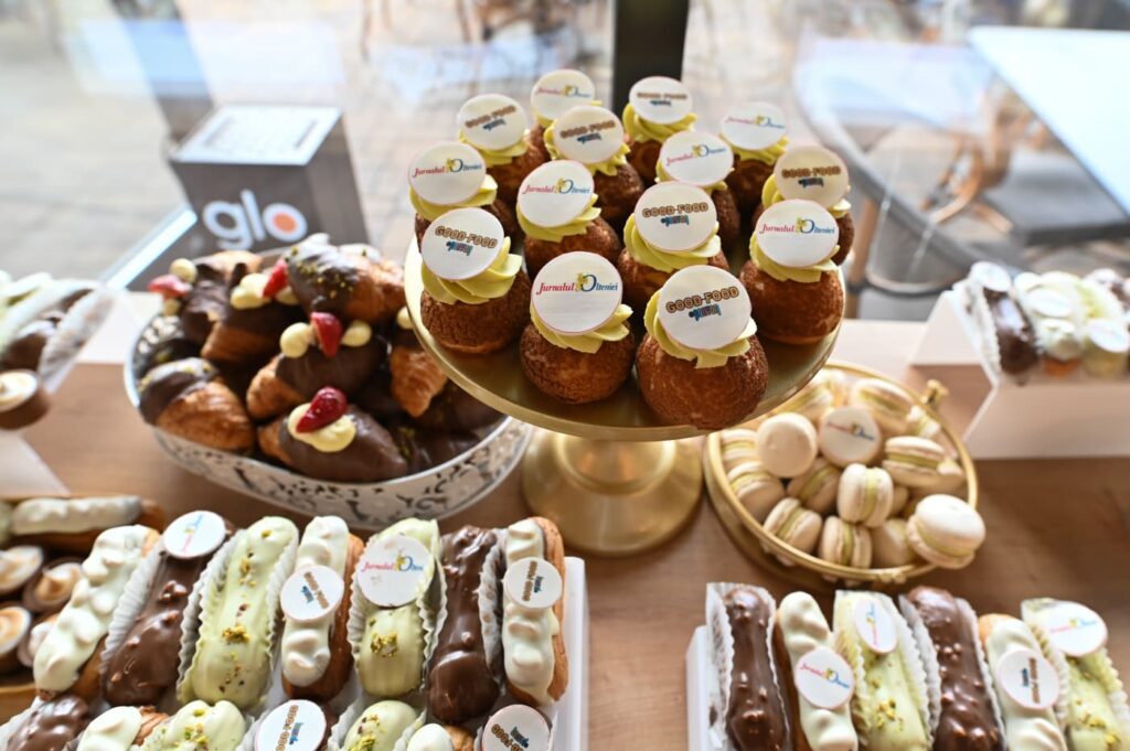 Bakery display with a gold tiered stand of cream puffs topped with round logo toppers, surrounded by macarons and chocolate pastries on a wooden table.