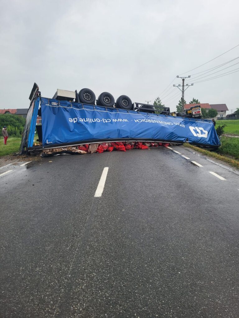 Overturned blue truck trailer blocking a two-lane road, wheels up, with red cargo bags spilled underneath.