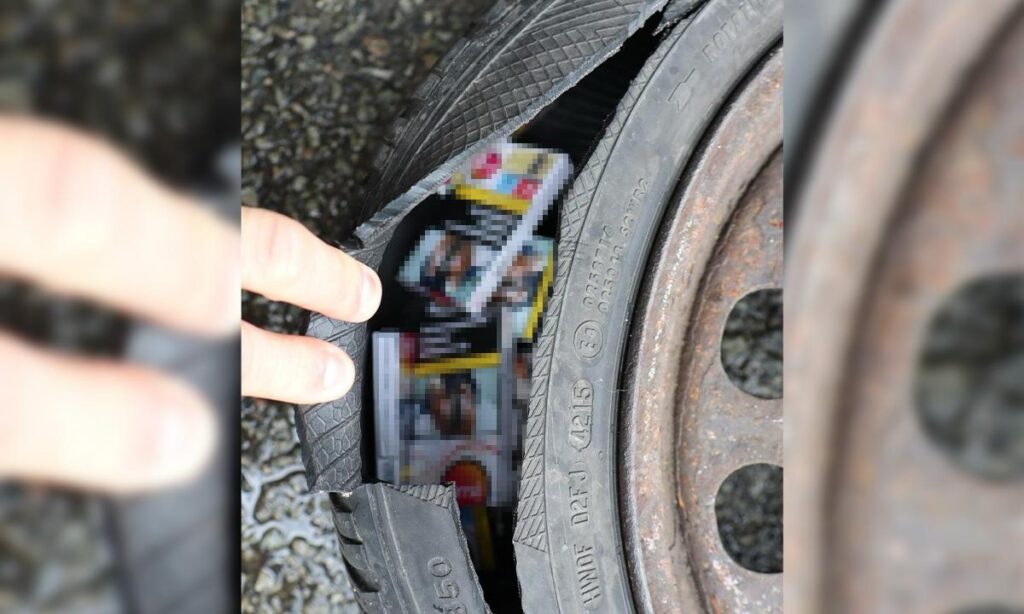 Close-up of a cracked tire with a rusted wheel rim; a small booklet or magazine is wedged into a hole in the tire, held by fingers.