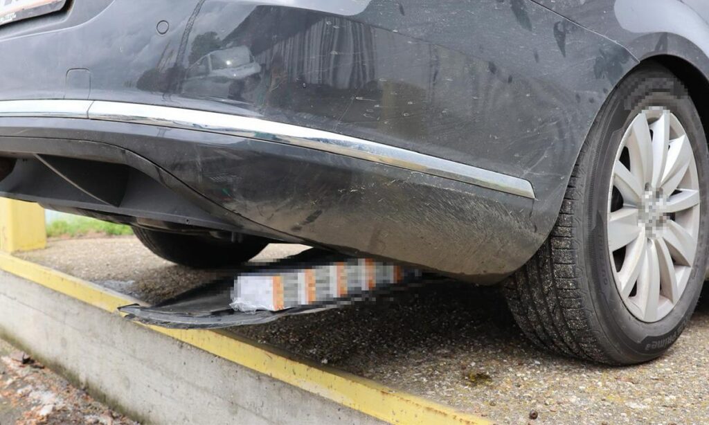 Gray sedan perched on a curb with the front bumper resting on a damaged package underneath it