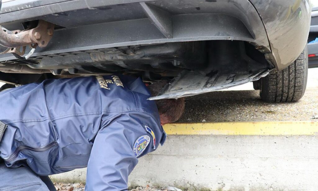 Person in a blue uniform lies under a car, inspecting the undercarriage near a curb (work activity). #informative image