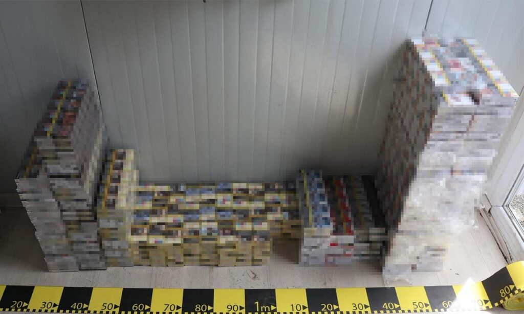 Stacks of VHS tapes arranged to form a wall-like outline in a corner, with a yellow-black measuring tape on the floor in the foreground of a gray room.