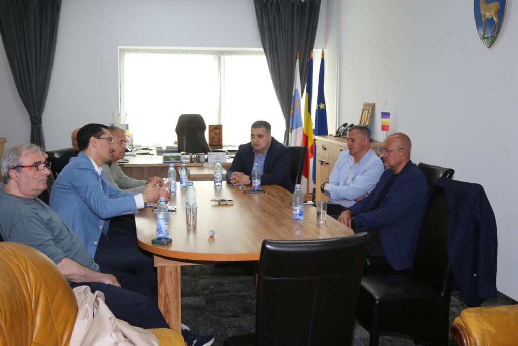 Group of men seated around a wooden conference table in a formal office meeting, flags in the background.