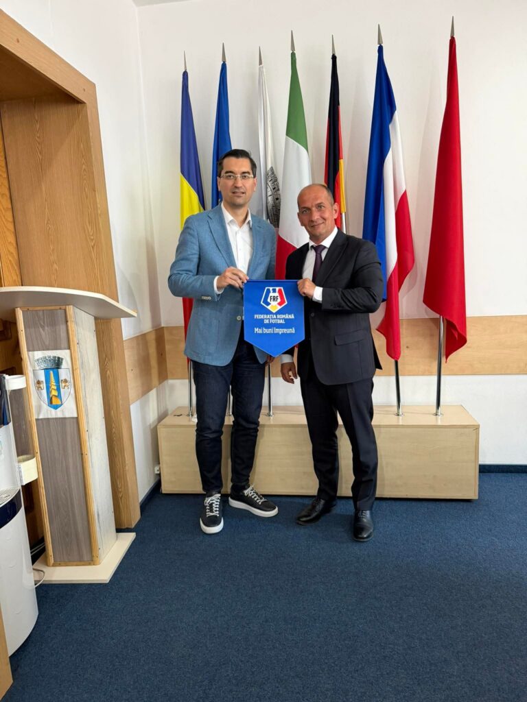 Two men in a formal indoor setting pose with a blue FRF banner, standing in front of a row of national flags behind them.