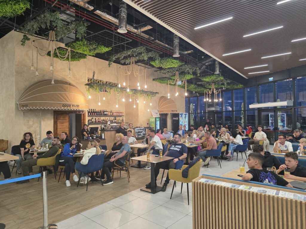 Busy indoor food court with many customers seated at tables under hanging light bulbs and greenery over head.