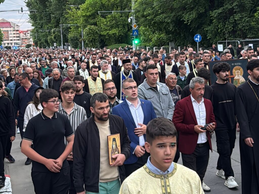 WhatsApp Image 2026-05-21 at 164746 - Jurnalul Olteniei – Știri din Craiova și Oltenia Large crowd of people, including clergy in ceremonial robes, marching down a city street; a priest near the front holds a religious icon while others carry icons or banners.