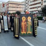 Orthodox priests in colorful ceremonial robes marching down a city street, carrying a large icon of two saints in the foreground.