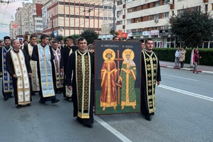 Orthodox priests in colorful ceremonial robes marching down a city street, carrying a large icon of two saints in the foreground.