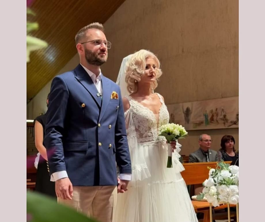 Bride in a white lace gown holds a bouquet beside a groom in a navy blazer at a church wedding, with guests seated in the background.