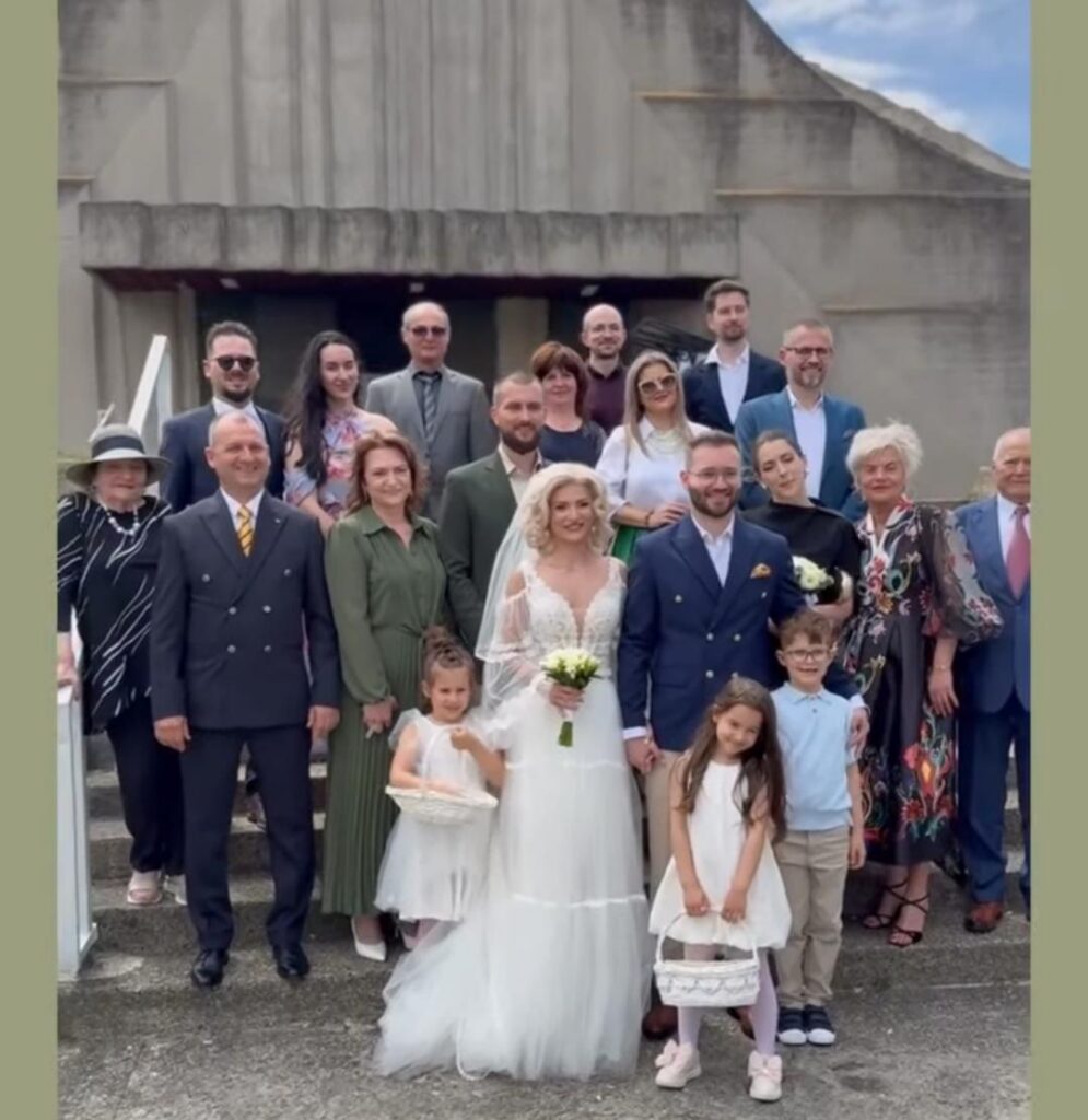 Bride and groom stand center in a wedding photo with guests and children posing on concrete steps outside a building; bride in a white gown holding a bouquet, groom in a navy suit.