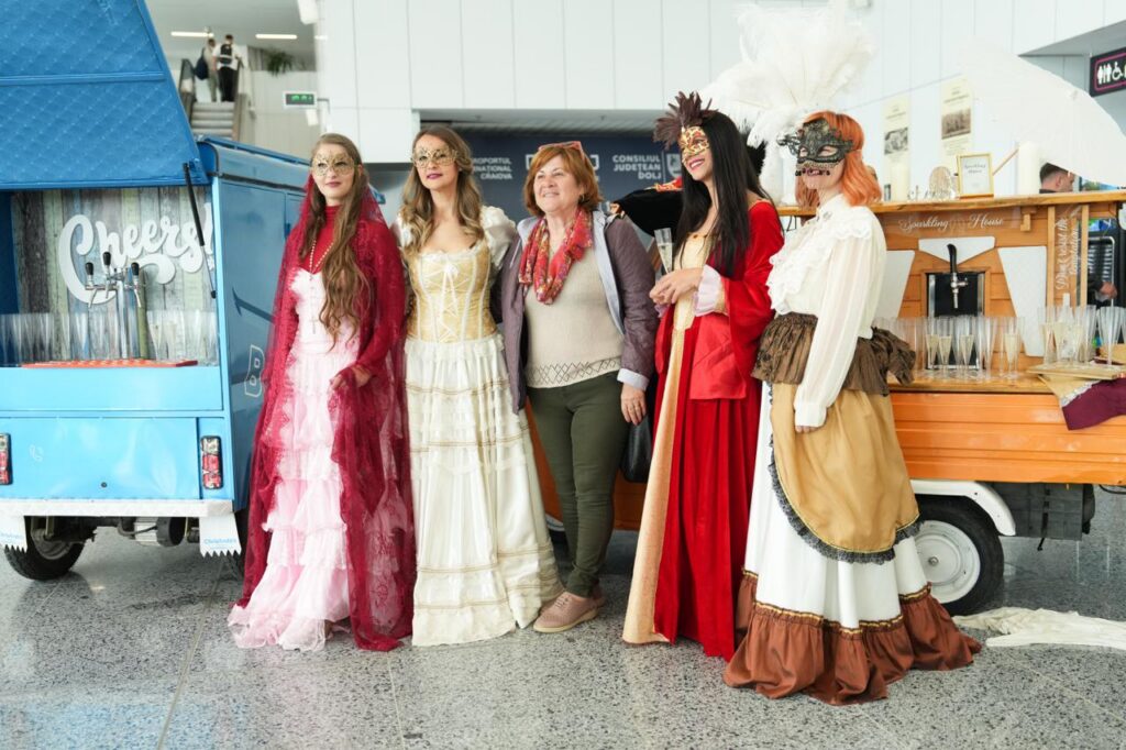 Group of five women in elaborate masquerade costumes and masks posing together indoors beside a blue service truck and an orange bar setup.