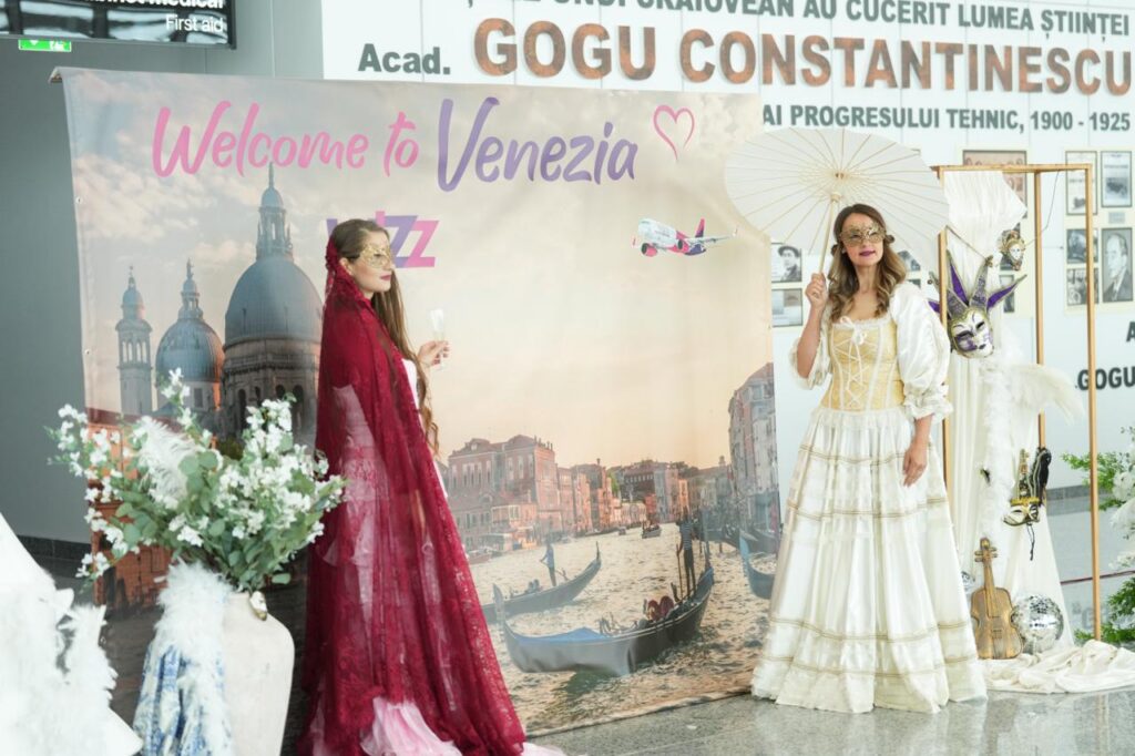 Two women in elaborate costumes pose in front of a 'Welcome to Venezia' backdrop with a gondola-themed scene and theatrical masks nearby.