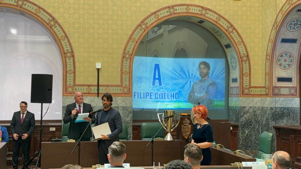 WhatsApp Image 2026-05-23 at 161347 1 - Jurnalul Olteniei – Știri din Craiova și Oltenia Formal ceremony in a historic, arched room; a man at a podium speaks into a microphone while another person holds papers, with a blue banner reading 'A FILIPE COELHO' behind them.